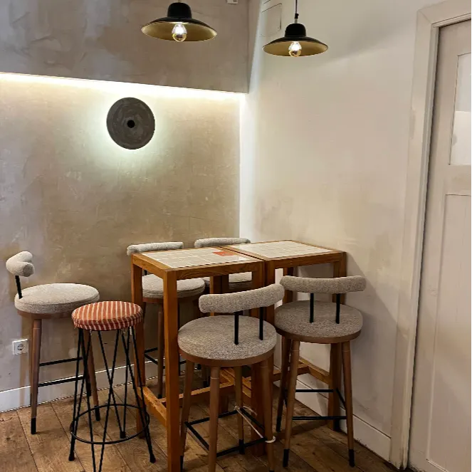 Rincón tranquilo de Maison Kayser Fernando VI con sillas altas, mesa de madera y lámparas colgantes en un espacio de paredes neutras.