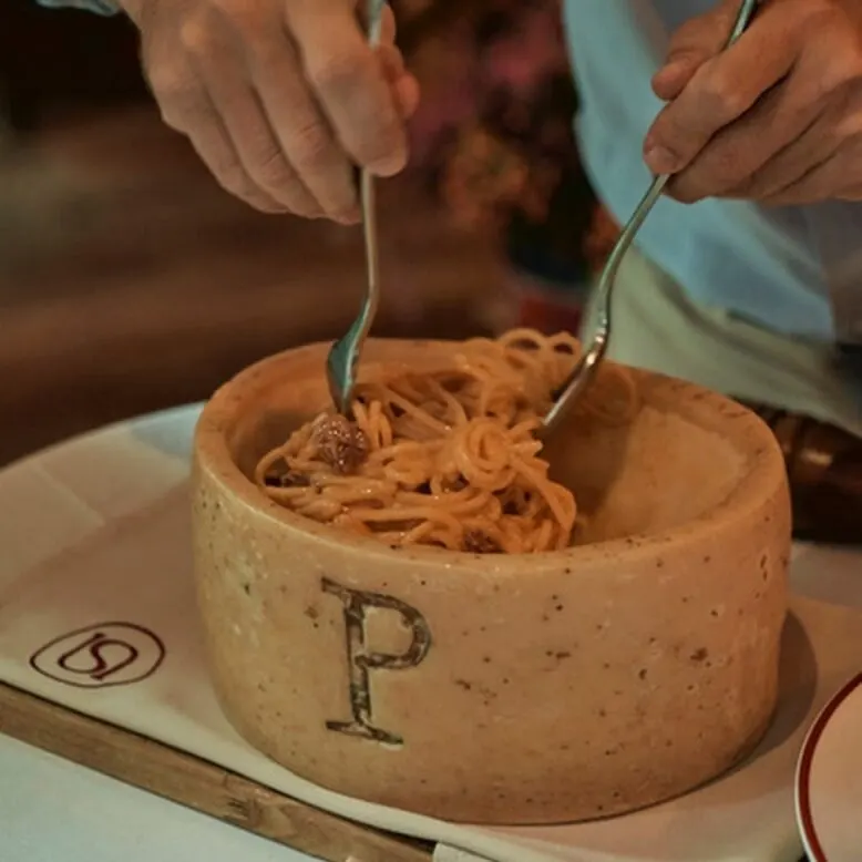 Persona mezcla espaguetis dentro de una rueda de queso con letra “P”, sobre mantel blanco y servilletas con logo ovalado con “S”.