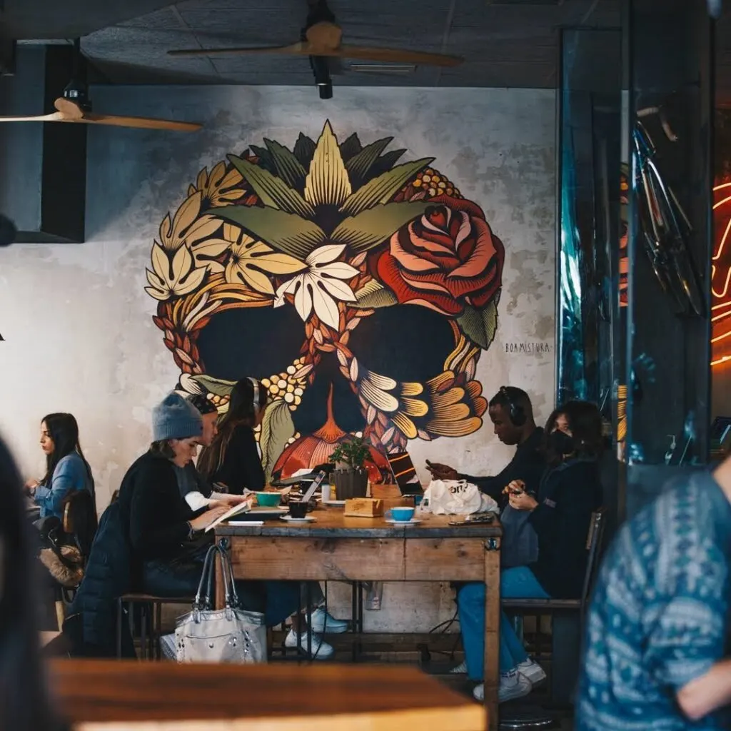 Grupo de personas en mesa de madera. Fondo con mural de calavera floral colorida. Ambiente relajado con ventiladores de techo visibles.