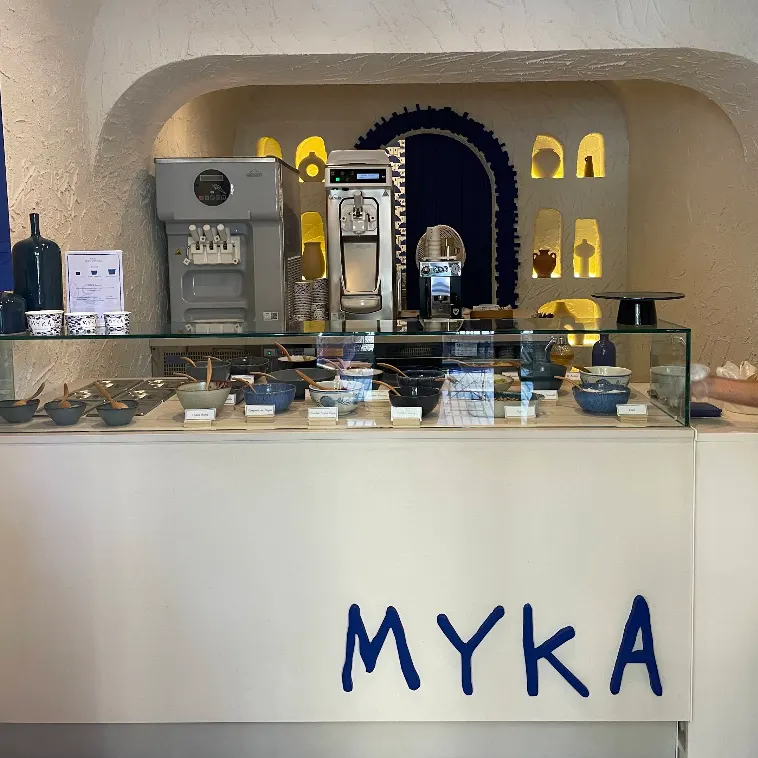 Mostrador de Myka Ortega y Gasset con vitrinas de toppings y fondo blanco con hornacinas iluminadas y puerta azul intensa.
