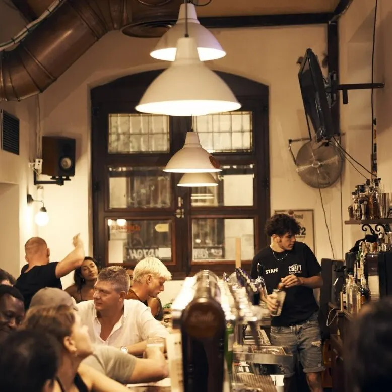 Interior de bar con clientes en la barra, bartender sirviendo bebidas, luces colgantes y ventanas grandes al fondo.