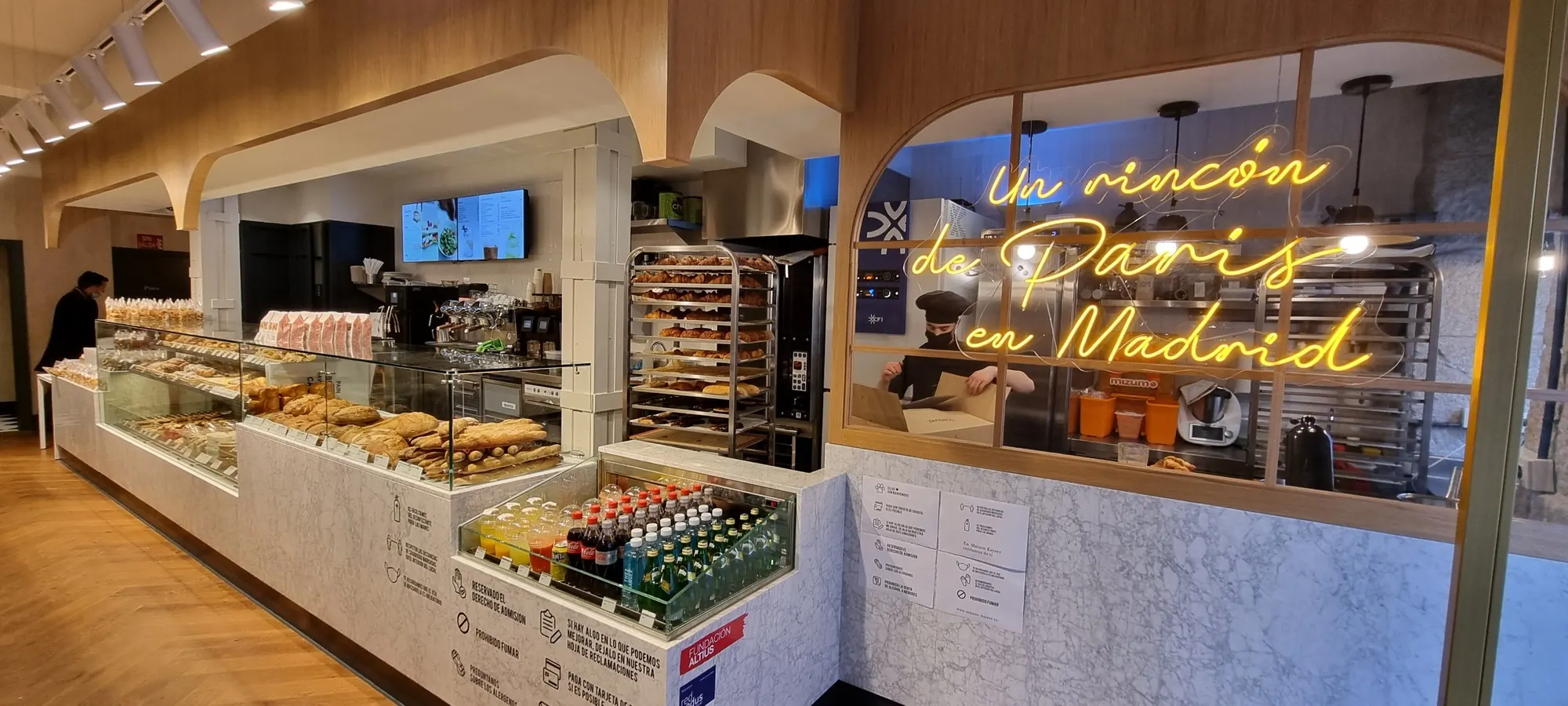 Mostrador lleno de bollería y bebidas junto a un obrador acristalado con neón que dice "Un rincón de París en Madrid" en Maison Kayser.