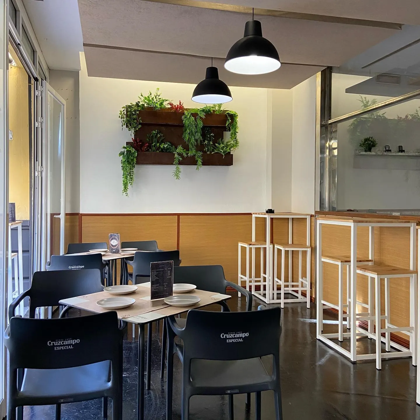 El Remate Bar y Tapas ofrece un espacio moderno con mesas de madera, sillas negras, plantas colgantes y luz cálida para una experiencia acogedora.