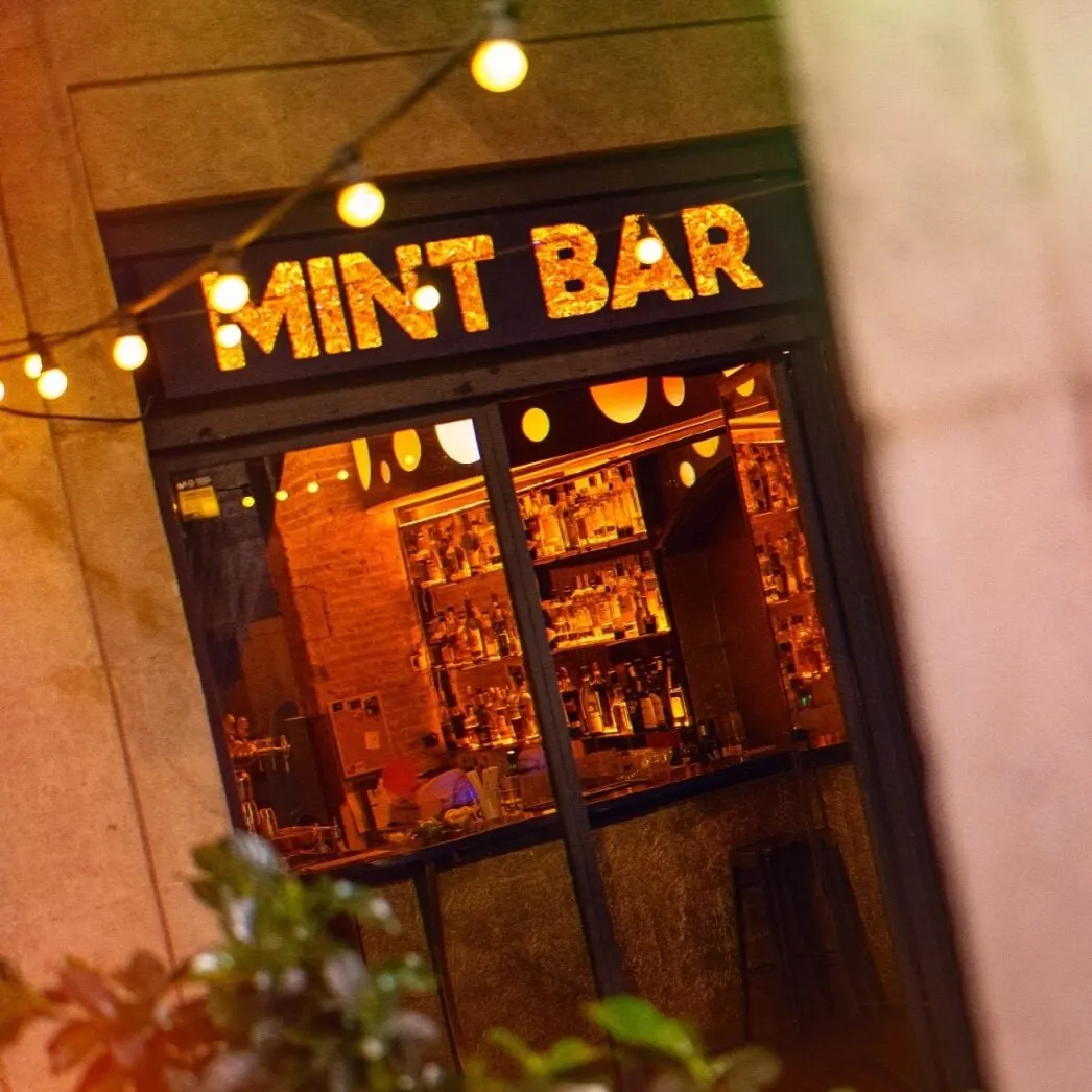 Entrada al MINT BAR con letrero iluminado, luces colgantes y ambiente acogedor; interior con estanterías llenas de botellas.