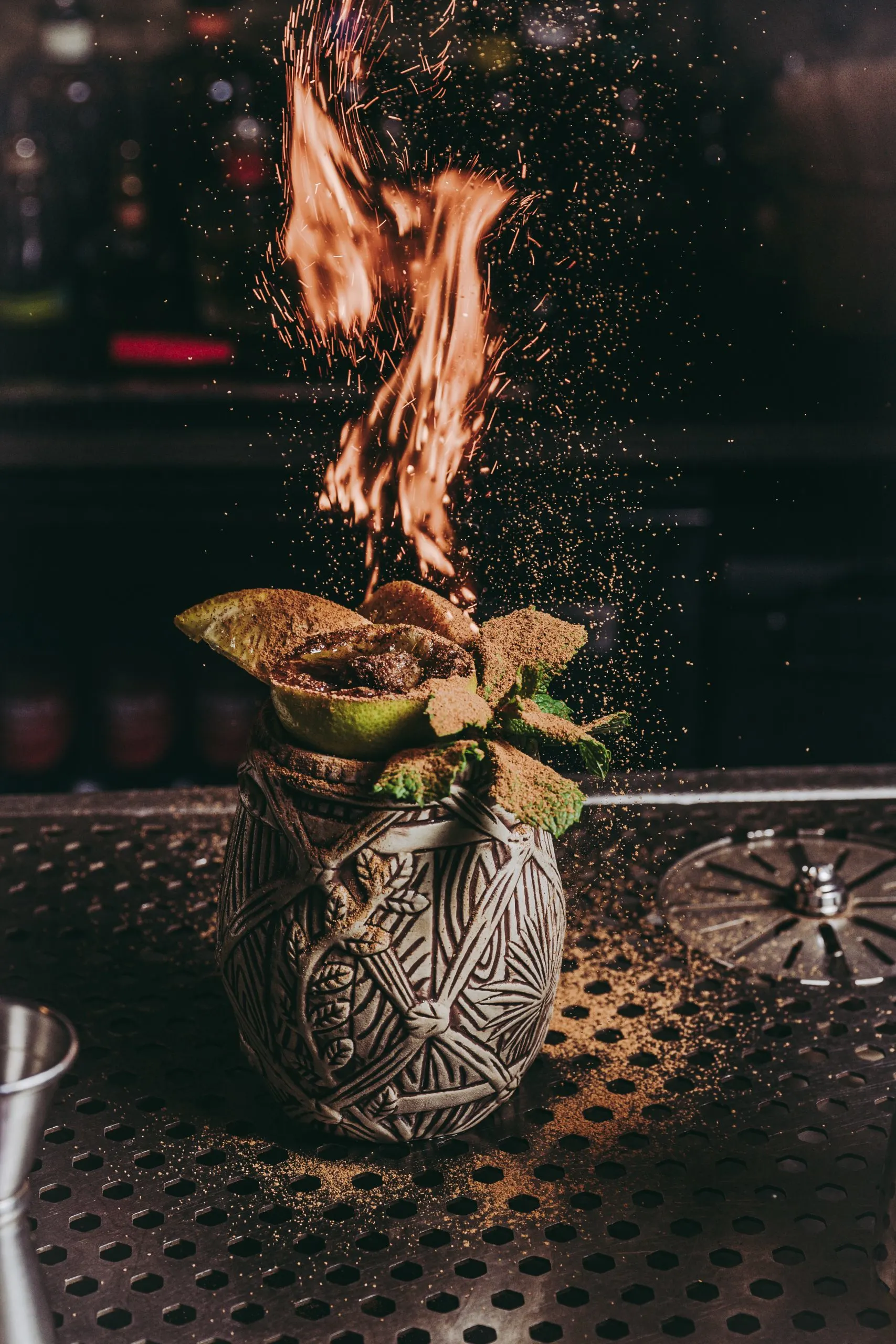 Cóctel en vaso artesanal con diseño tribal, decorado con cítricos y menta, acompañado de llamas y chispas que realzan su presentación.