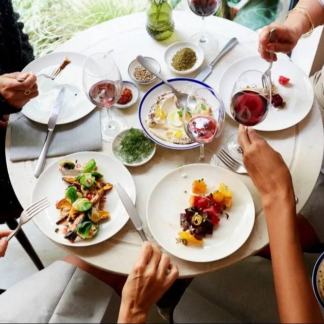 Café interior con mesas redondas, sillas de madera, plantas decorativas y luz suave en ambiente moderno y acogedor.
