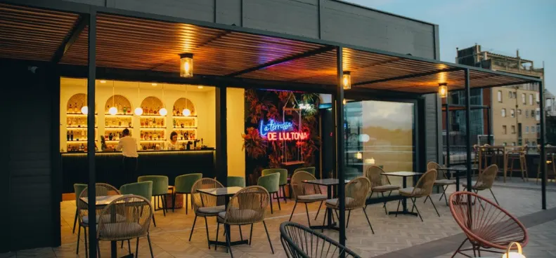 Terraza moderna con bar iluminado al fondo, sillas y mesas vacías al aire libre y luces cálidas al atardecer.