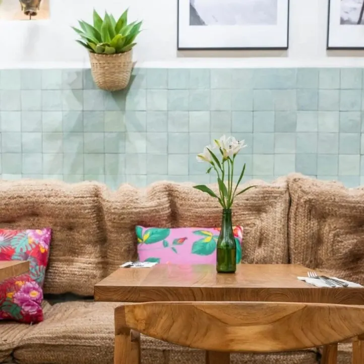 Rincón de café con mesa y silla de madera, cojines marrón, almohadas coloridas, flores blancas, pared azul con fotos y planta colgante.