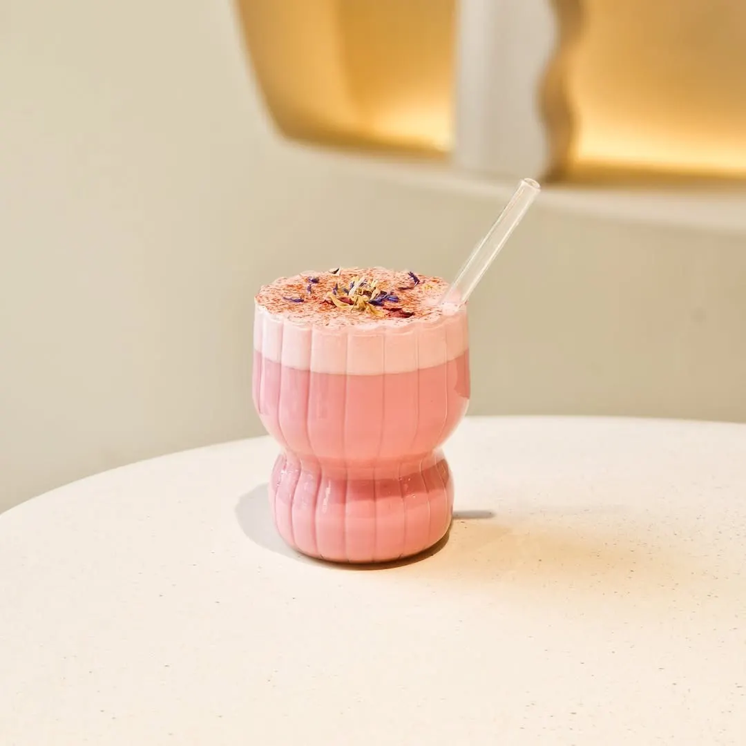 Bebida rosa servida en un vaso curvo a juego con espuma decorada con pétalos y pajita transparente sobre una mesa blanca de estilo minimalista.