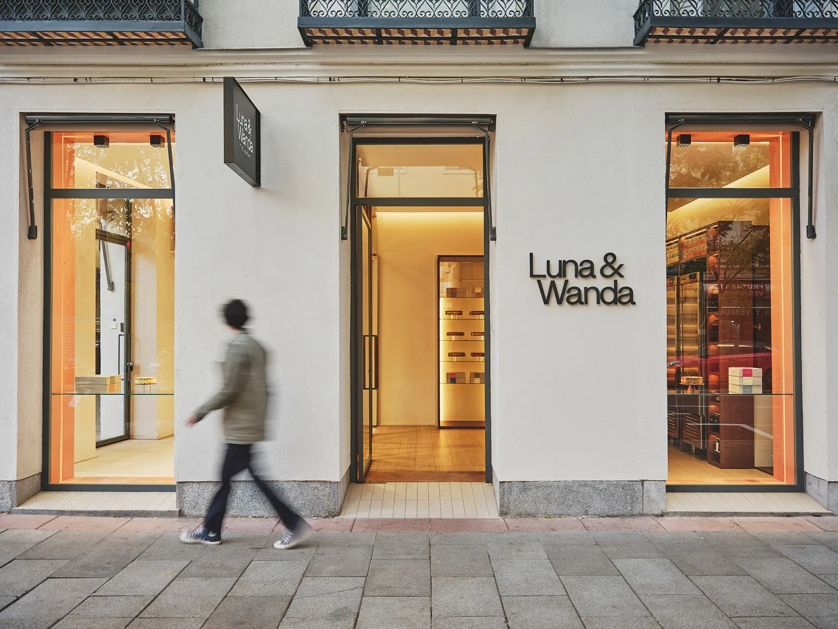 Fachada minimalista de Luna & Wanda Ferraz con grandes ventanales, luz cálida interior y rótulo negro sobre pared blanca.