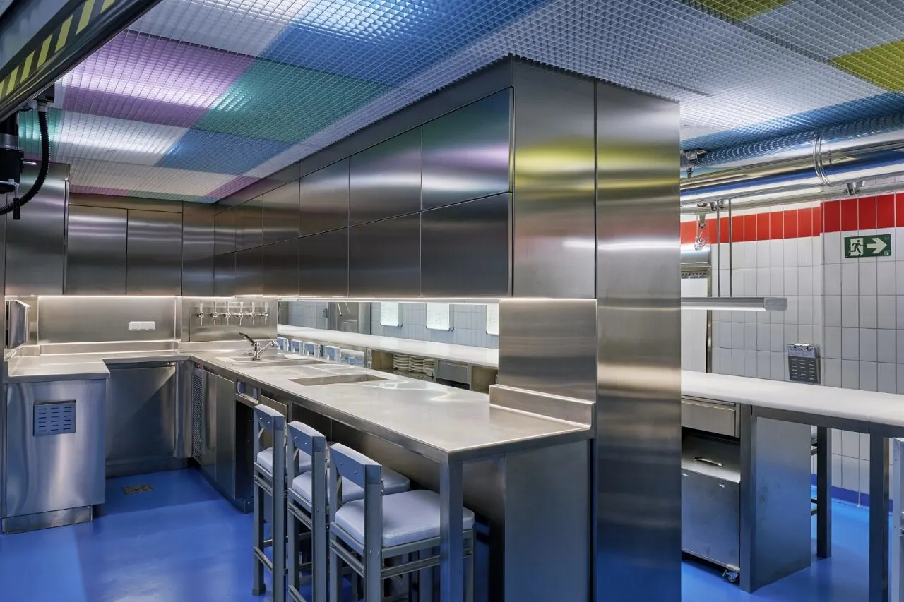 Cocina vista de Sala de Despiece con barra de acero inoxidable, taburetes metálicos, azulejos blancos y techo de rejilla con paneles de colores.