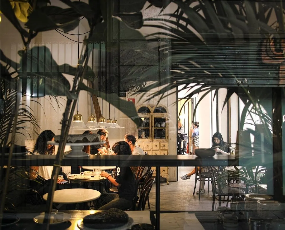 Café acogedor visto desde una ventana, con gente conversando y comiendo. Reflejos de plantas e interior crean un ambiente cálido y social.