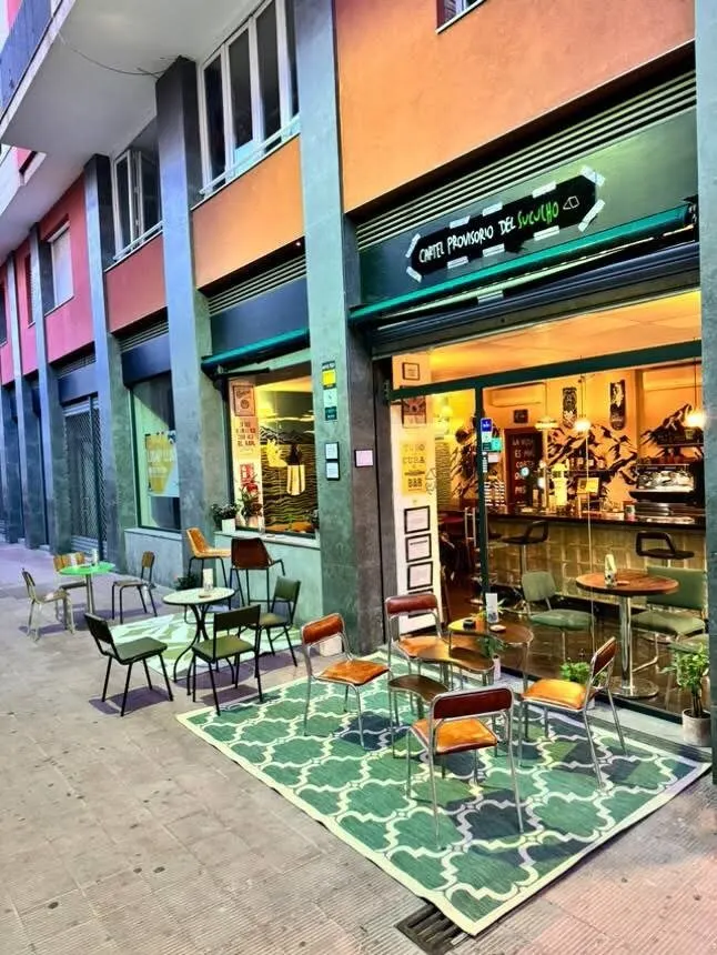 Exterior de “Café Provisorio de Sucucho” con terraza sobre alfombra verde, mesas y sillas. Interior acogedor visible tras ventanas grandes.
