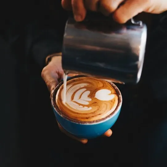 Persona vertiendo leche en café para crear arte latte con formas de corazones y hojas; taza azul sostenida con una mano.
