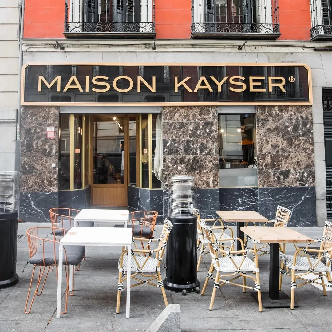 Fachada de Maison Kayser con sillas trenzadas y mesas al aire libre frente a una pared de mármol oscuro y una fachada rojiza elegante.