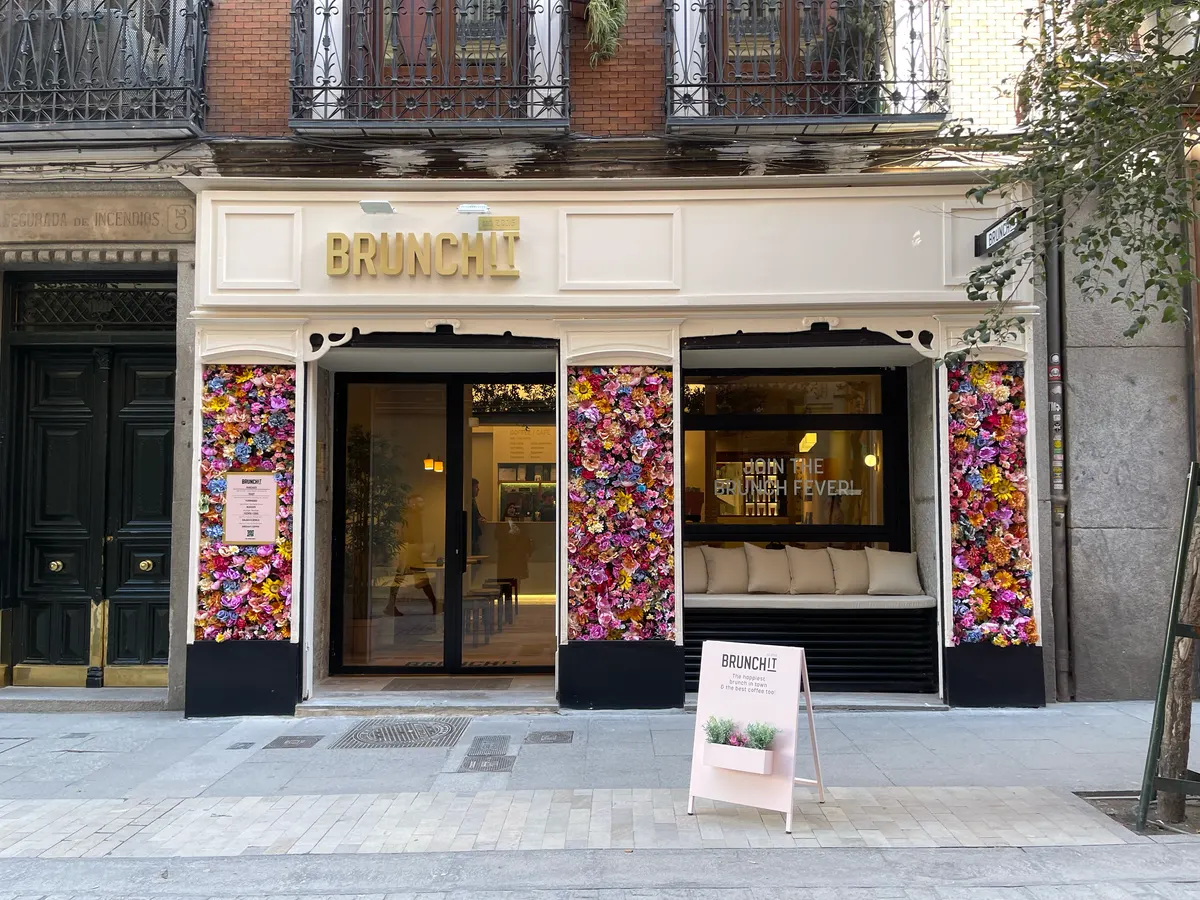 Fachada de Brunchit Alhóndiga con marco de flores de colores, letrero dorado, cartel promocional y banco con cojines frente al ventanal.