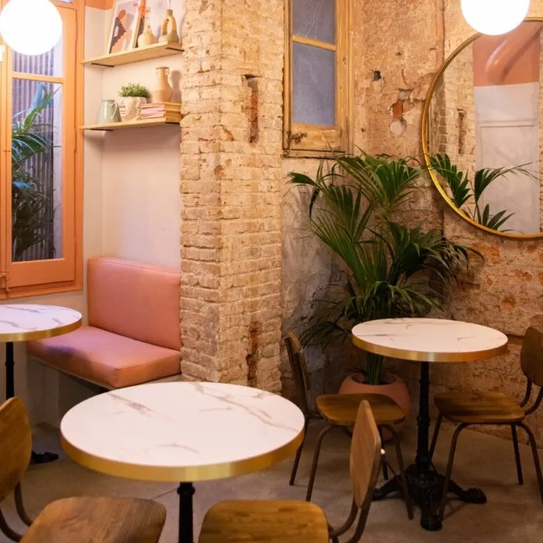 Café acogedor con mesas redondas, pared de ladrillo, espejo circular, luces colgantes, banco rosa y estantería con libros y plantas.