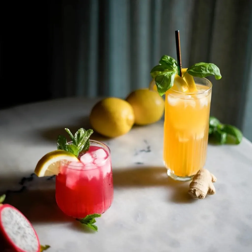 Cócteles rojo y naranja con limón, menta y albahaca sobre mármol, junto a limones, jengibre y pitahaya partida.