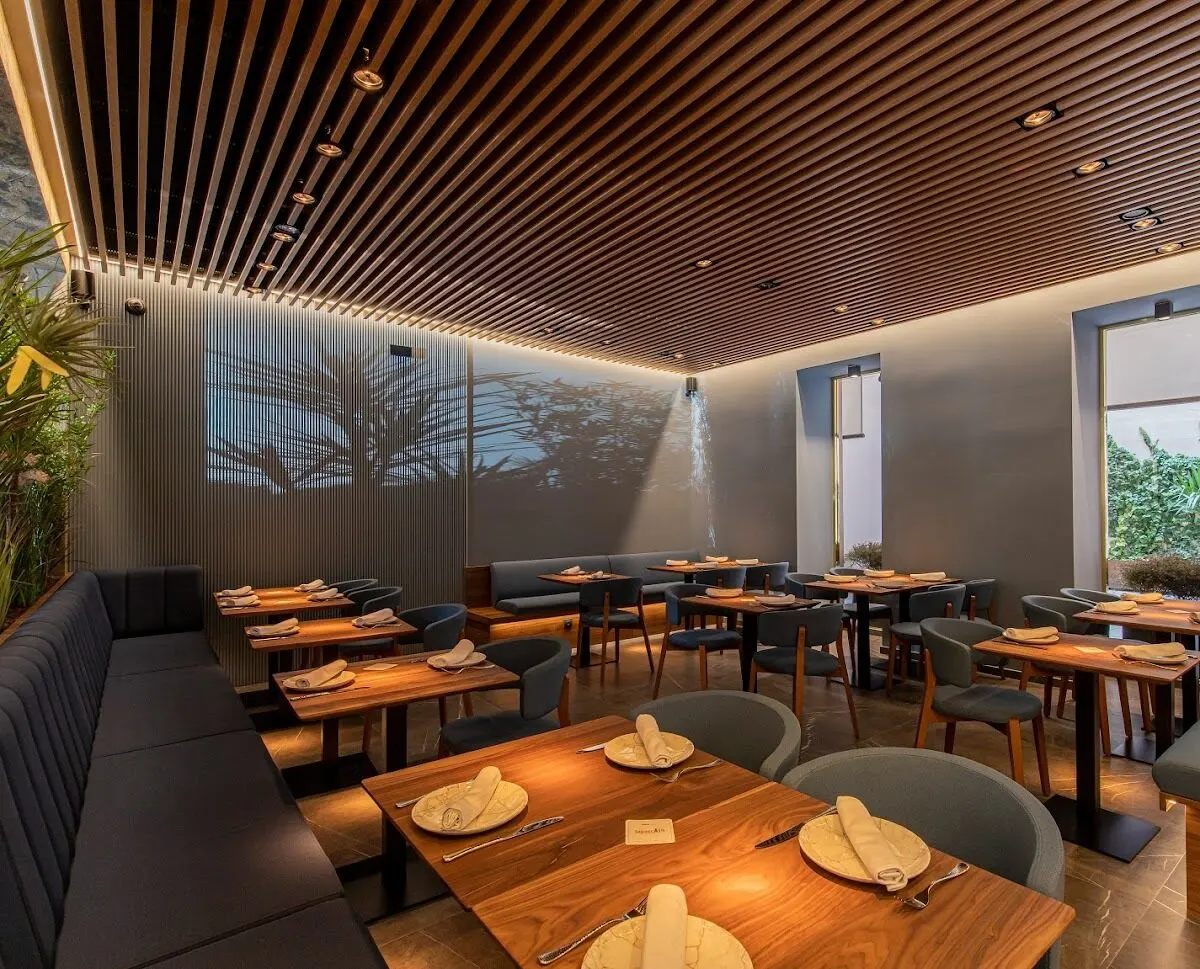Restaurante moderno con mesas de madera, luz natural, techo con listones y plantas decorativas. Ambiente luminoso y bien organizado.