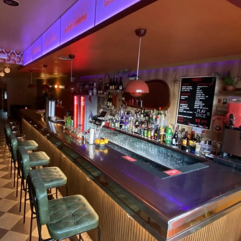 Bar con barra larga, taburetes verdes, botellas en estanterías, luces colgantes y letreros luminosos con frases como “good times”.