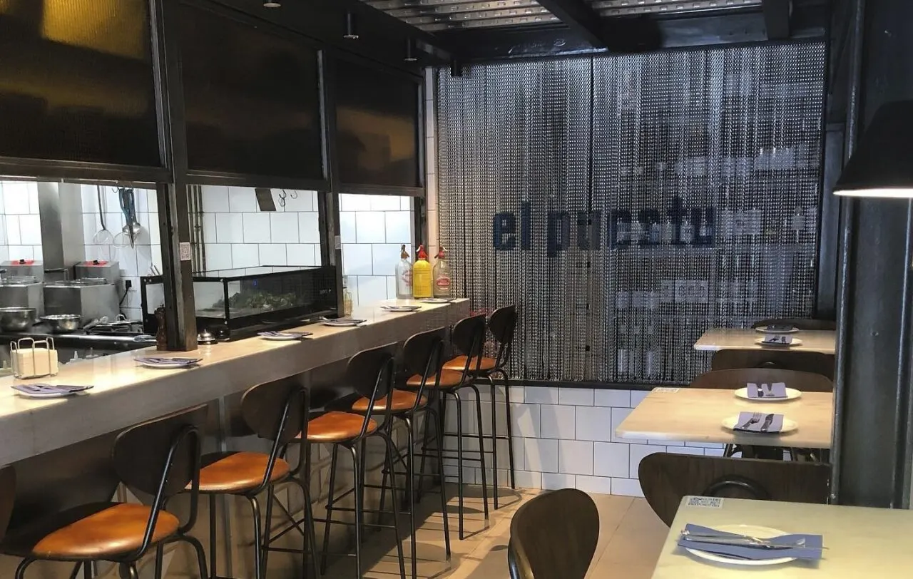 Restaurante de estilo moderno e industrial con cocina abierta, barra con sillas altas y mesas puestas. Fondo con cortina metálica y texto “el puestu".