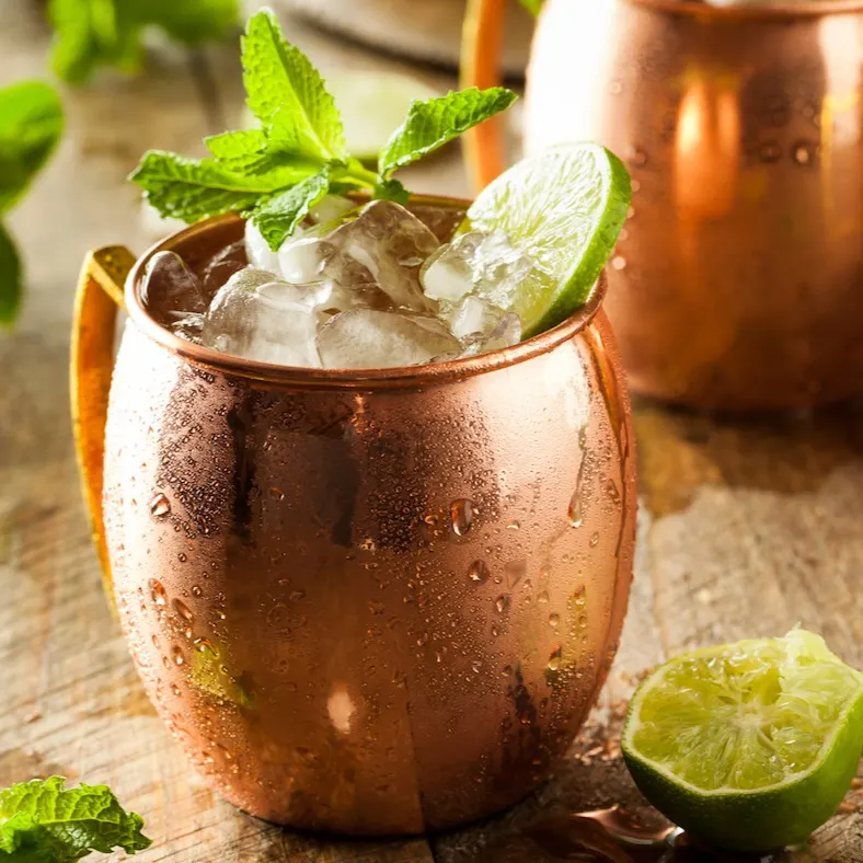 Copa de cobre con cóctel, hielo picado, menta y lima, sobre mesa rústica con gotas de agua, hojas sueltas y cítricos cortados.