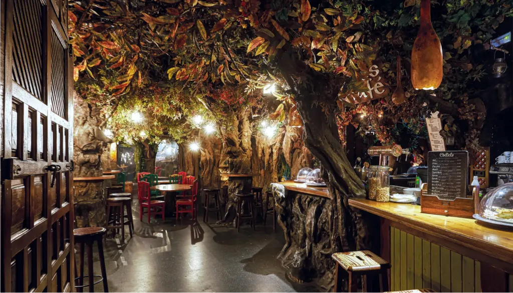 Cafetería temática con decoración de bosque: follaje artificial, mesas coloridas y barra estilo tronco. Iluminación cálida y ambiente envolvente.