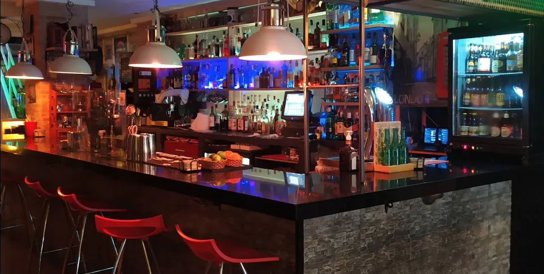 La barra del bar London con taburetes rojos, lámparas colgantes y estanterías repletas de botellas iluminadas en tonos azules y cálidos.