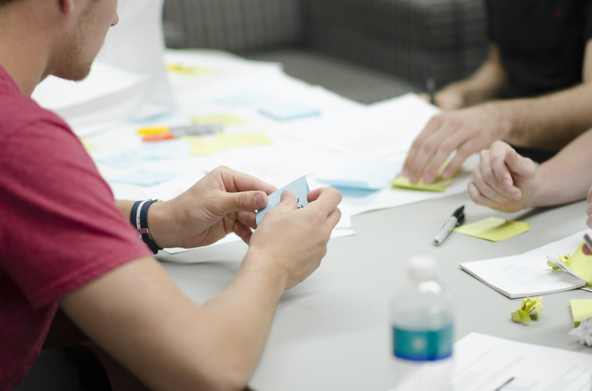 A picture of a team working around a table littered with paper, post it notes, and other materials.