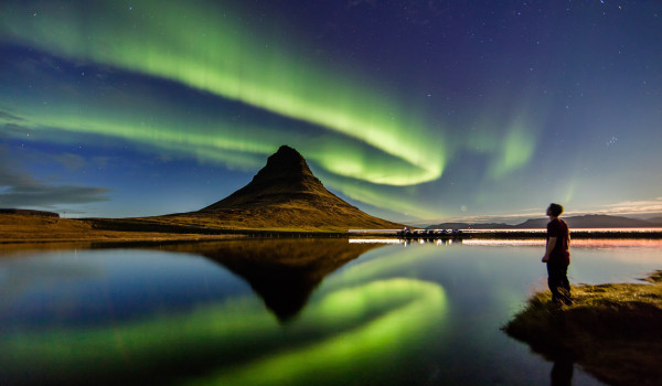 Person standing near a reflective lake with a cone-shaped mountain and green aurora borealis overhead.