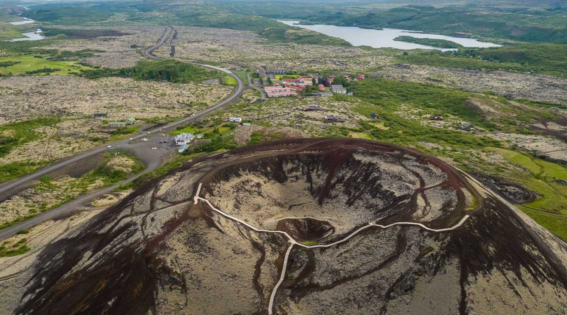 An aerial view of a picturesque countryside village in Iceland showcases a red-roofed church, modern buildings, and lush green fields surrounded by rolling hills under a partly cloudy sky.