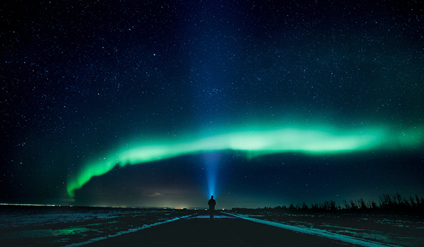  Person standing on a road, shining a light into the sky under an arching green aurora borealis.