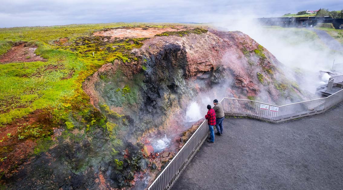 Two visitors stand by a metal railing, observing steaming geothermal vents emerging from a moss-covered volcanic hillside, creating an otherworldly landscape in Iceland’s geothermal region.