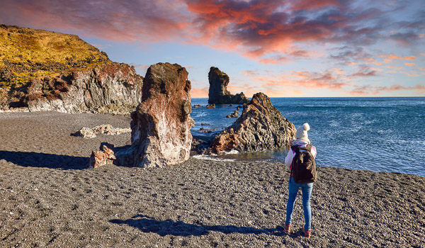 Person standing on Djúpalónssandur beach with black sand, unique rock formations, and a vibrant sunset sky.