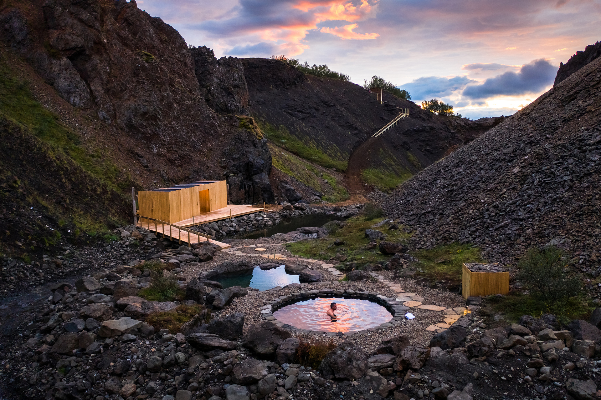 A secluded geothermal hot spring, surrounded by rugged volcanic hills, features a single bather enjoying the warm waters at sunset, with wooden walkways and minimalistic structures blending into the natural landscape.