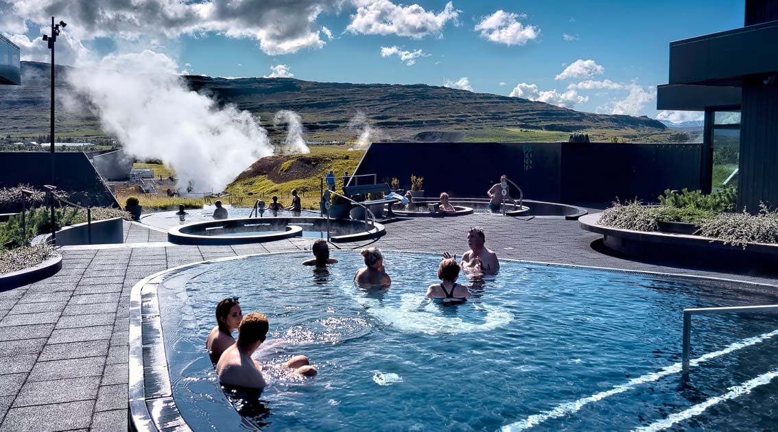 Krauma Spa in Iceland features geothermal hot pools with visitors relaxing in the warm waters, surrounded by modern architecture and a scenic backdrop of rolling hills and rising steam from natural hot springs.