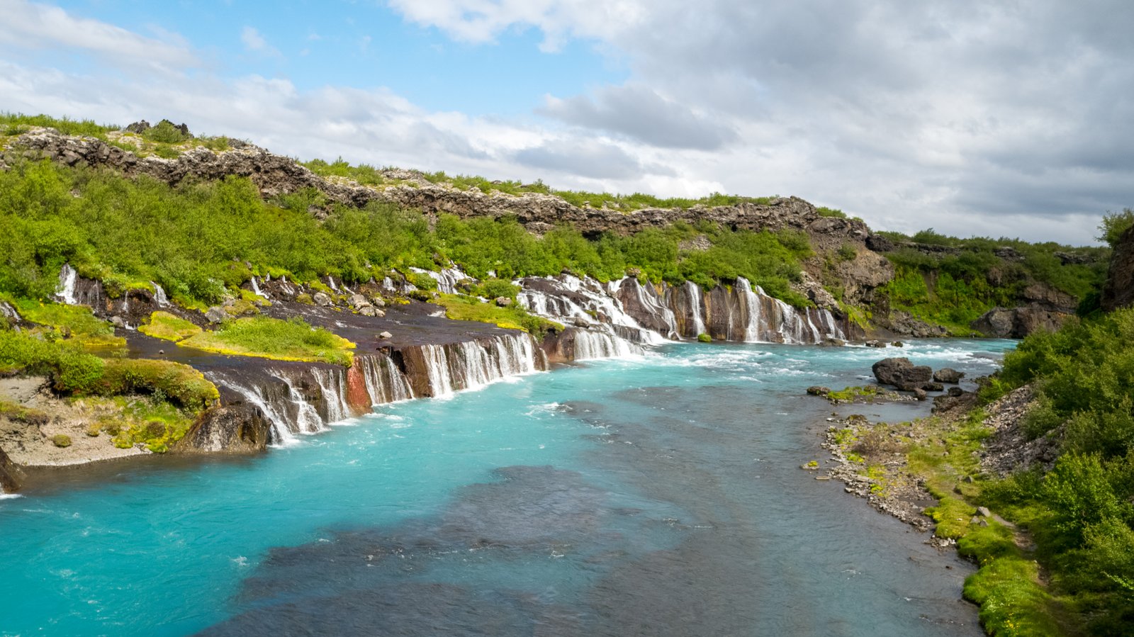 Hraunfossar, a series of stunning waterfalls in Iceland, flows over lava rock formations into a vibrant turquoise river, surrounded by lush greenery and distant mountains.