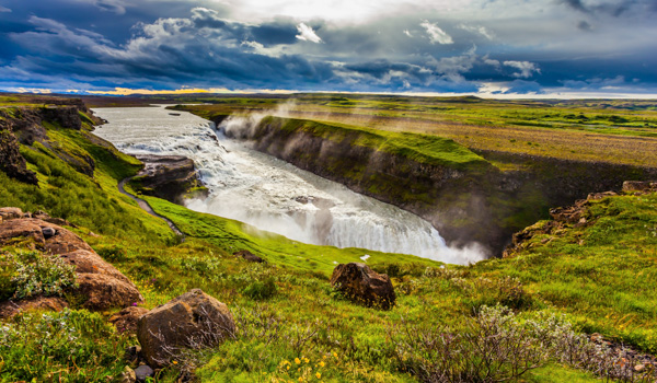 Majestic view of Gullfoss waterfall cascading into a rugged canyon, with lush green landscape and dramatic sky in the background.