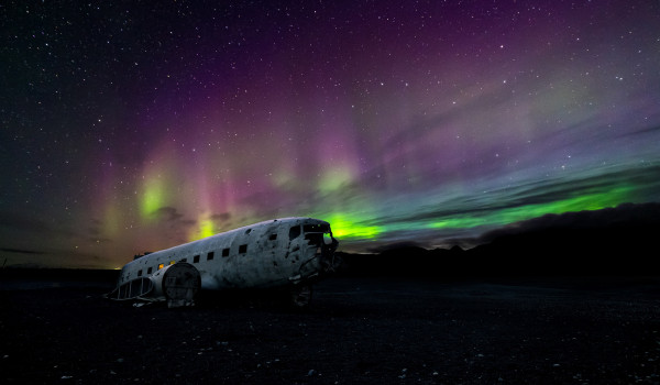 Abandoned airplane wreckage under a vibrant purple and green aurora borealis in a starry night sky.