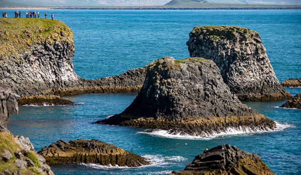 Rock formations and cliffs on the coastline between Arnarstapi Hellnar, surrounded by blue ocean water and a grassy cliff edge with people.