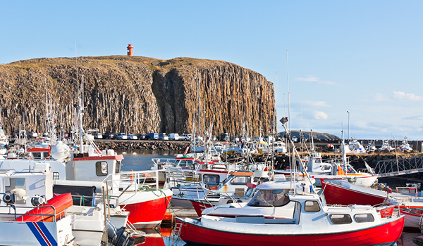 Boats docked at Stykkishólmur harbor with a rocky cliff and a small lighthouse in the background.