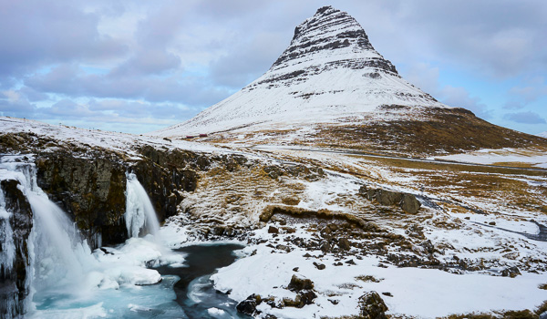 Snow-covered Kirkjufell mountain with a partially frozen Kirkjufellsfoss waterfall in the foreground under a cloudy sky in Iceland.