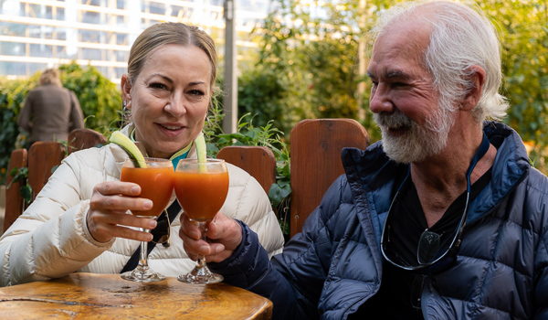 Smiling couple toasting with tomato drinks at Fridheimar greenhouse, surrounded by lush green plants.