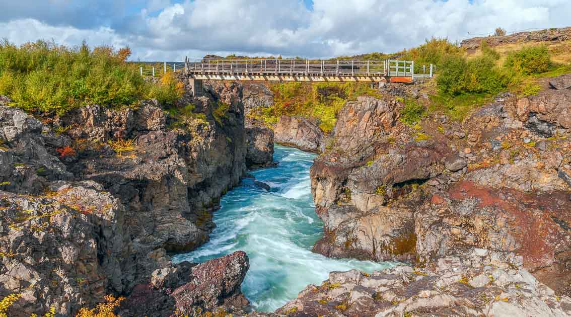 Barnafoss, a fast-flowing glacial river in Iceland, rushes through a rugged volcanic canyon beneath a wooden bridge, surrounded by rocky cliffs and lush greenery under a partly cloudy sky.