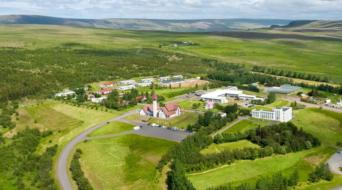 An aerial view of a picturesque countryside village in Iceland showcases a red-roofed church, modern buildings, and lush green fields surrounded by rolling hills under a partly cloudy sky.