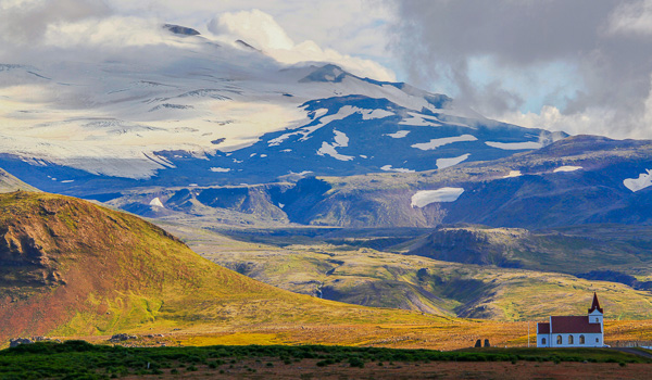 Small church in the foreground with Snæfellsjökull glacier and surrounding green mountains under a bright sky.