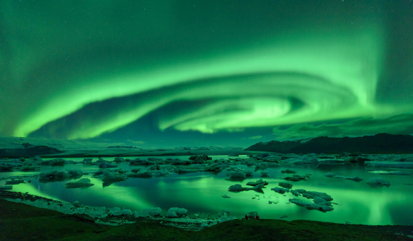 Swirling green aurora borealis over a lagoon with floating icebergs and distant mountains.