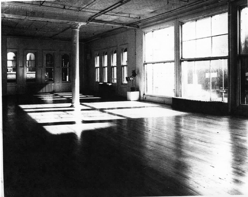 A black and white photograph of the interior of The Kitchen's Soho loft space in 1975. Daylight is coming through the large windows on the right. There are no people visible in the space.