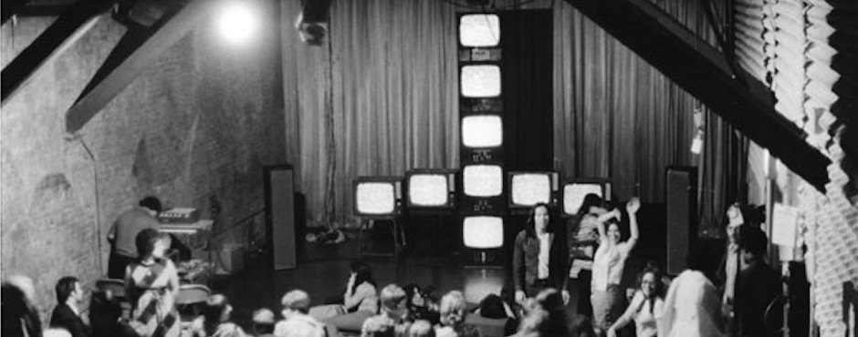 A black and white, archival photo from the original Kitchen showing a video installation of stacked television monitors and people gathered around socializing.