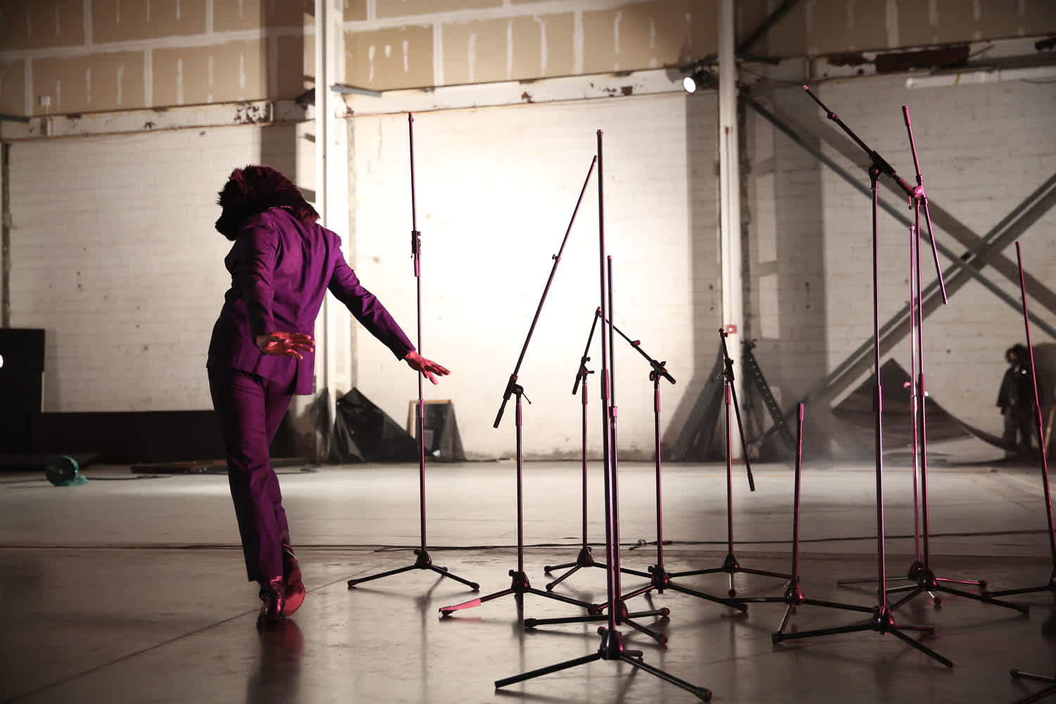 In a warehouse space, a figure in a purple suit and wearing a wolf mask faces away from the camera with their arms reaching behind them. To the right, there is a group of mic stands. 